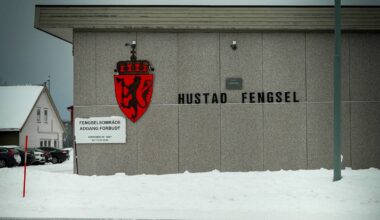 Hustad Fengsel er avbildet med en grå fasade og et rødt skjold med en løve. Det er snødekket bakken foran bygget, og et skilt varsler om at området er avgrenset. I bakgrunnen vises en liten, hvit bolig. Skyene er mørke, noe som gir et trist inntrykk av omgivelsene. (Bildebeskrivelsen er laget av en KI-tjeneste)