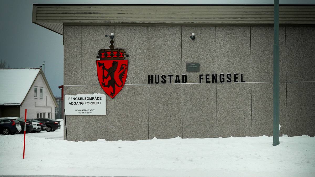 Hustad Fengsel er avbildet med en grå fasade og et rødt skjold med en løve. Det er snødekket bakken foran bygget, og et skilt varsler om at området er avgrenset. I bakgrunnen vises en liten, hvit bolig. Skyene er mørke, noe som gir et trist inntrykk av omgivelsene. (Bildebeskrivelsen er laget av en KI-tjeneste)
