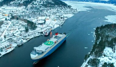 Et stort frakteskip ligger i en smal elv omgitt av snødekte hus og trær. Området er dekket av snø, med isbiter i vannet. Fjellene i bakgrunnen understreker vinterlandskapet. Det er klart vær, og lysene fra bygningene er synlige.