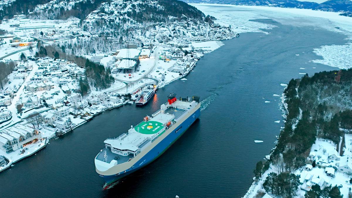Et stort frakteskip ligger i en smal elv omgitt av snødekte hus og trær. Området er dekket av snø, med isbiter i vannet. Fjellene i bakgrunnen understreker vinterlandskapet. Det er klart vær, og lysene fra bygningene er synlige.