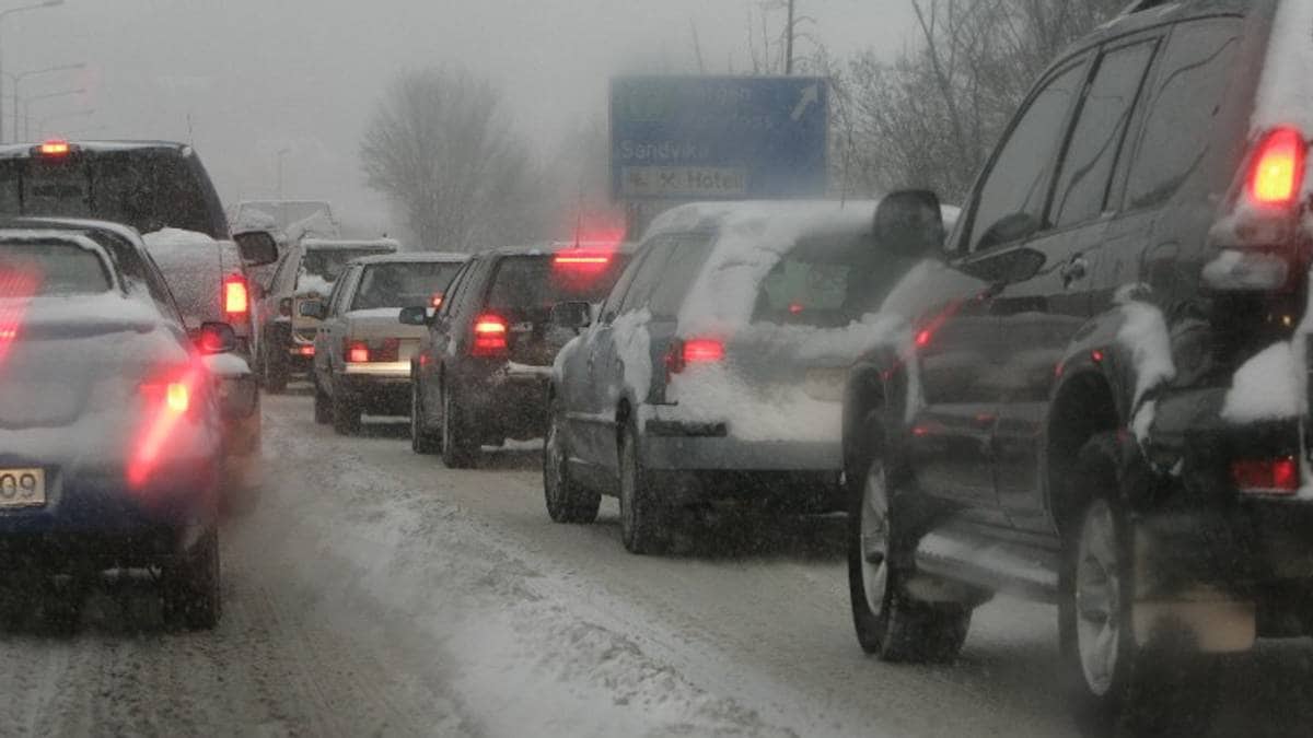 Trafikk står stille i snøvær, med flere biler som ligger tett i en rekke. Veibanen er dekket av snø, noe som skaper vanskelige kjøreforhold. I bakgrunnen er det veiskilt som viser retning mot Sandvika. Omgivelsene er preget av snø og lav sikt. (Bildebeskrivelsen er laget av en KI-tjeneste)
