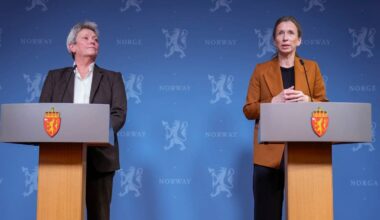 To personer står bak hver sin talerstol med mikrofon under en pressekonferanse i Norge. Bakgrunnen er dekorert med riksvåpenet til Norge og teksten "NORWAY". Talerstolene har embetssegl preget på fremsiden. (Bildebeskrivelsen er laget av en KI-tjeneste)