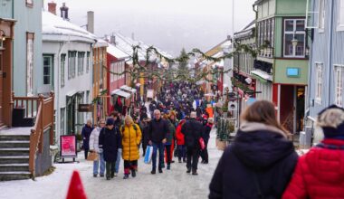 En travel gate i en by med folk som går forbi fargerike bygninger, dekorert for vinteren. Flere personer bærer shoppingposer og det er snø på bakken. Julepynt og granbar henger over gaten, som antyder en festlig atmosfære. Stemningen er livlig med mange besøkende i området. (Bildebeskrivelsen er laget av en KI-tjeneste)