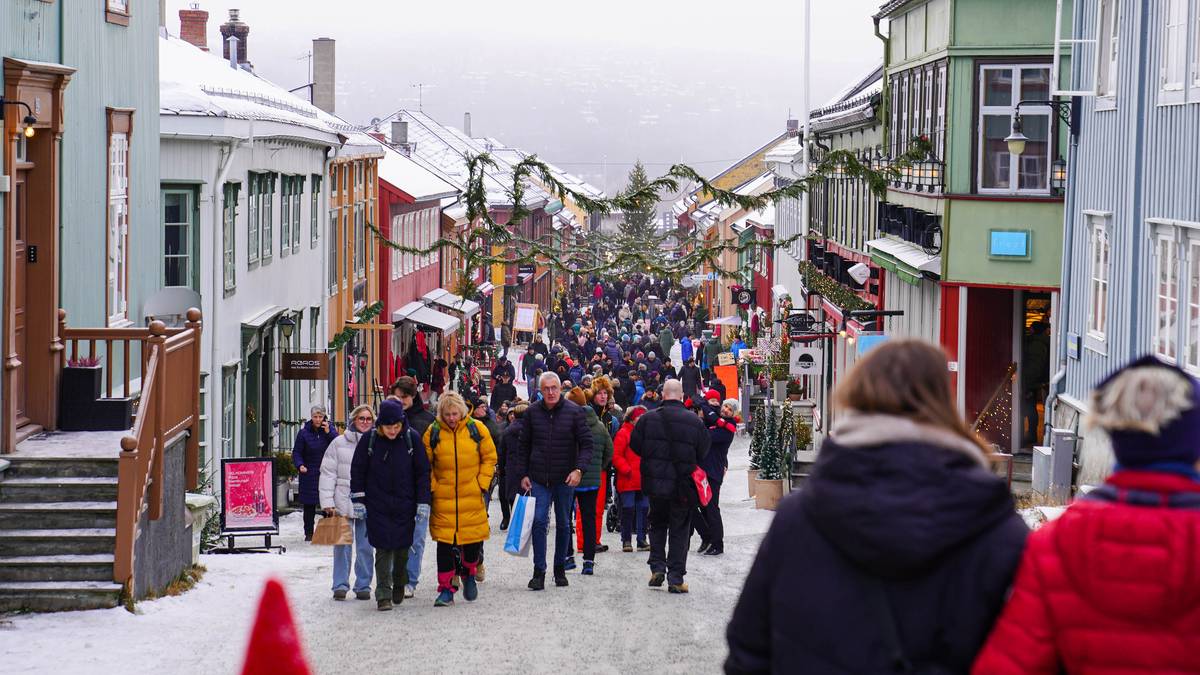 En travel gate i en by med folk som går forbi fargerike bygninger, dekorert for vinteren. Flere personer bærer shoppingposer og det er snø på bakken. Julepynt og granbar henger over gaten, som antyder en festlig atmosfære. Stemningen er livlig med mange besøkende i området. (Bildebeskrivelsen er laget av en KI-tjeneste)
