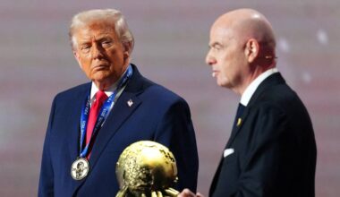 En person i dress med medalje står ved siden av en annen person i mørk dress. Den første holder en statuett av en globe. Begge ser mot hverandre i en seremoniell setting. Omgivelsene er svakt opplyst med en dekorativ bakgrunn. (Bildebeskrivelsen er laget av en KI-tjeneste)