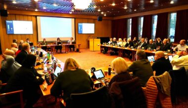 Et møte finner sted i et rom med en lang bordoppsett, hvor mange deltakere sitter. Noen personer ser på skjermer, mens andre er i samtale. Det er presentasjoner på veggene, og flere deltakere bruker bærbare datamaskiner. Vinduer med rød gardin gir naturlig lys til rommet. (Bildebeskrivelsen er laget av en KI-tjeneste)