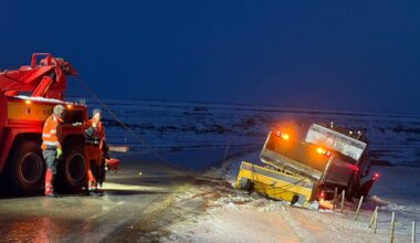 Tre menn i arbeidstøy står ved siden av en veltet lastebil på en glatte vei. En kranbil er plassert nær lastebilen, og mennene forbereder redningsoperasjonen. Området er dekket av snø, og det er mørkt, med lys fra kranbilen og blinkende varsellamper. Veien er smal med et rekkverk på siden, og det ser ut til å være glatte forhold. (Bildebeskrivelsen er laget av en KI-tjeneste)