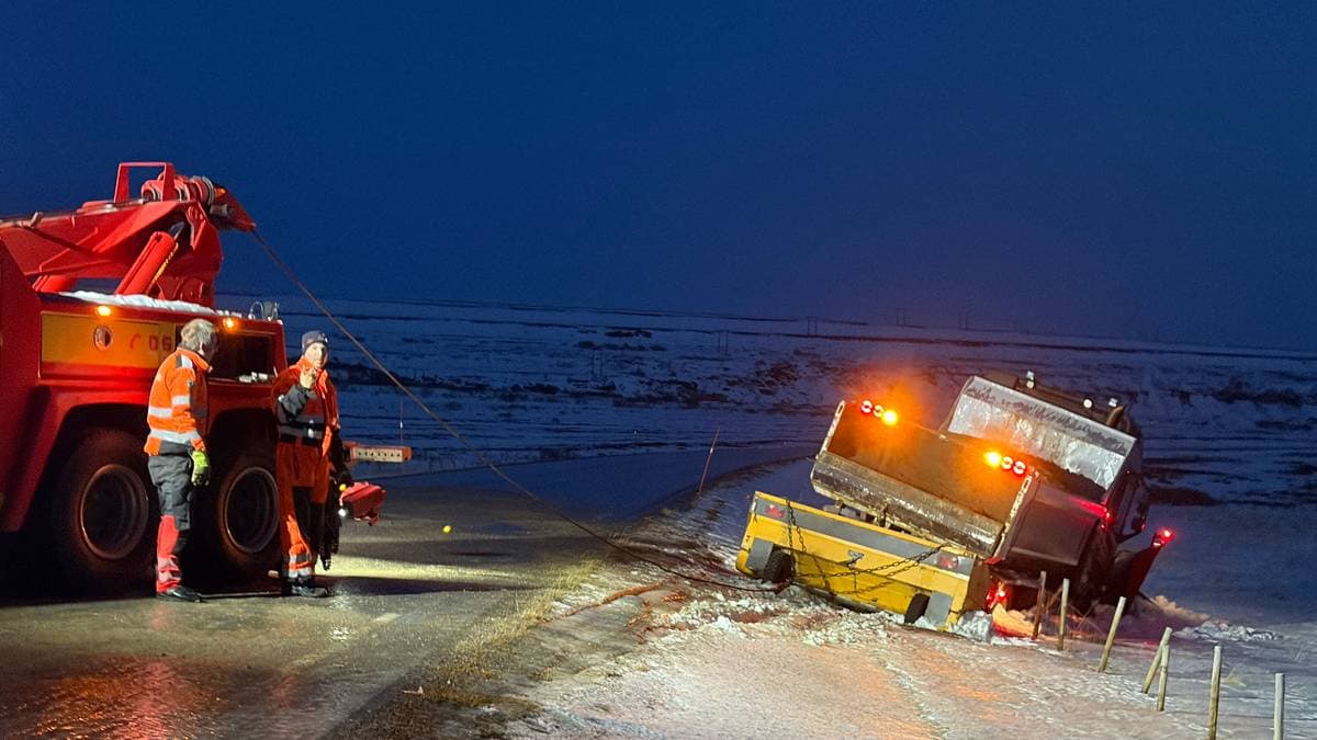 Tre menn i arbeidstøy står ved siden av en veltet lastebil på en glatte vei. En kranbil er plassert nær lastebilen, og mennene forbereder redningsoperasjonen. Området er dekket av snø, og det er mørkt, med lys fra kranbilen og blinkende varsellamper. Veien er smal med et rekkverk på siden, og det ser ut til å være glatte forhold. (Bildebeskrivelsen er laget av en KI-tjeneste)