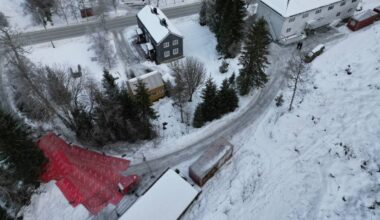 Oversiktsbilde av et snødekt boligområde med hus i forskjellige farger. En rød presenning ligger på bakken til venstre, mens to personer går langs veien. Veien svinger mot høyre i bildet, omringet av trær og snø. Det er en rolig vinterstemning i området. (Bildebeskrivelsen er laget av en KI-tjeneste)