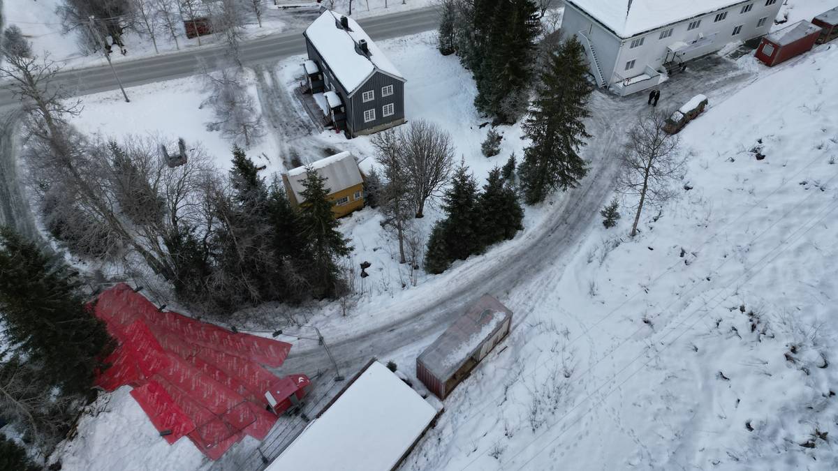 Oversiktsbilde av et snødekt boligområde med hus i forskjellige farger. En rød presenning ligger på bakken til venstre, mens to personer går langs veien. Veien svinger mot høyre i bildet, omringet av trær og snø. Det er en rolig vinterstemning i området. (Bildebeskrivelsen er laget av en KI-tjeneste)