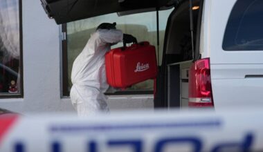 En person iført beskyttelsesdrakt bærer en rød koffert med "Leica" trykket på, inn i en bil med åpen luke. Kofferten ser ut til å inneholde utstyr, muligens relatert til undersøkelse eller fotografering. I bakgrunnen er det vinduer til en bygning, og det er synlig noe dekorasjon. Bildet gir inntrykk av en situasjon knyttet til profesjonelt arbeid eller oppdrag. (Bildebeskrivelsen er laget av en KI-tjeneste)