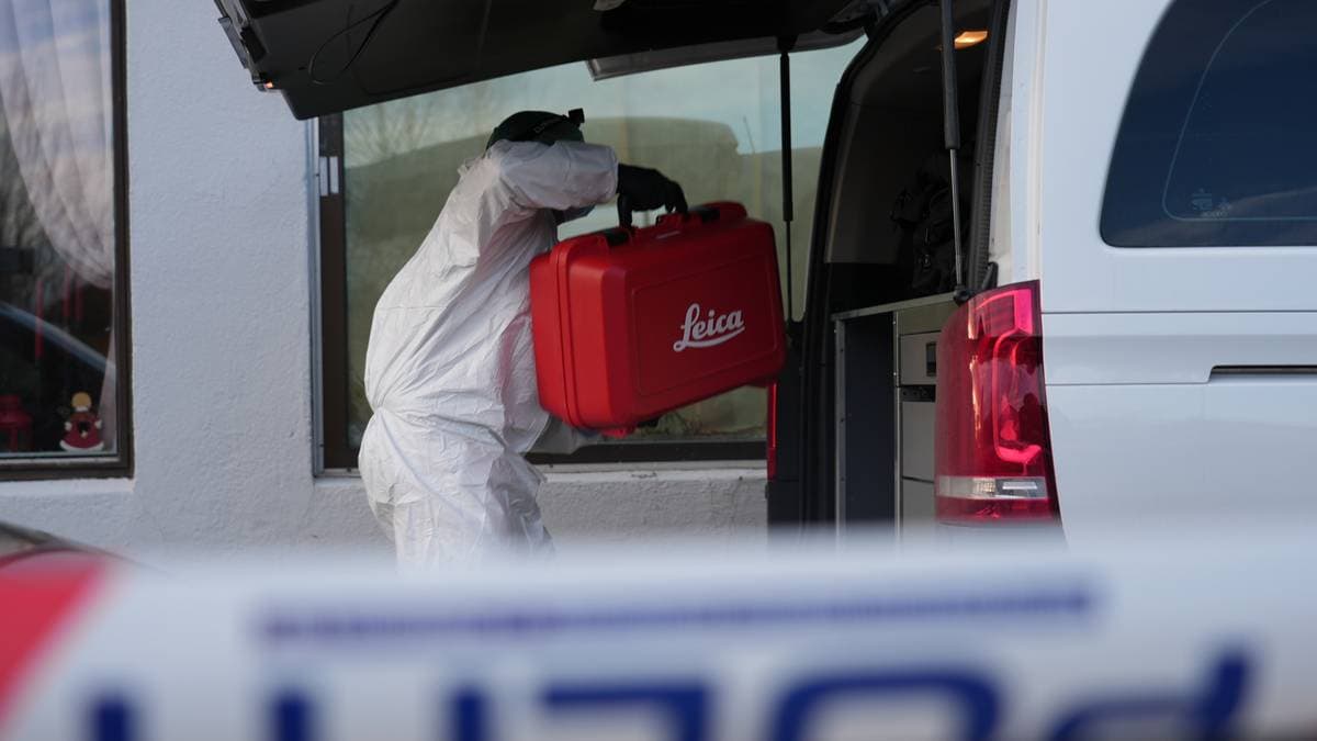 En person iført beskyttelsesdrakt bærer en rød koffert med "Leica" trykket på, inn i en bil med åpen luke. Kofferten ser ut til å inneholde utstyr, muligens relatert til undersøkelse eller fotografering. I bakgrunnen er det vinduer til en bygning, og det er synlig noe dekorasjon. Bildet gir inntrykk av en situasjon knyttet til profesjonelt arbeid eller oppdrag. (Bildebeskrivelsen er laget av en KI-tjeneste)