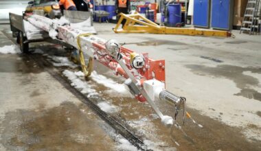 Bildet viser en snøfreser foran en bil i en verkstedlignende setting. En person i oransje arbeidsklær jobber med utstyret, mens en annen står bak bilen. Gulvet er dekket av snørester, og ulike verktøy og utstyr er synlig i bakgrunnen. Belysningen er kunstig, noe som gir en tydelig oversikt over arbeidsområdet. (Bildebeskrivelsen er laget av en KI-tjeneste)