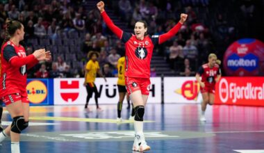 En spiller i rød drakt jubler etter et mål under en håndballkamp. Flere spillere fra begge lag er synlige i bakgrunnen. Det er en stor mengde publikum i arenaen. Reklamebannerne rundt banen bidrar til sportsmiljøet.