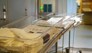 Bildet viser flere klare, plastkasser plassert på traller i et rom som sannsynligvis er en sykehusavdeling. Hver kasse inneholder stabler av berg med tekstiler og papirdokumenter, som kan indikere forberedelser for et medisinsk inngrep. Omgivelsene virker ryddige og profesjonelle, med en dør i bakgrunnen som gir tilgang til et annet rom. Det er et grønt lys i nærheten, som kan signalisere aktivitet eller observasjon i området. (Bildebeskrivelsen er laget av en KI-tjeneste)