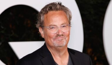 En mann i svart jakke og grå T-skjorte poserer foran en lys bakgrunn. Han har kort, bølgete hår. Bildet er tatt på et arrangement, med grønne planter synlige i bakgrunnen. Det er noe uklart i ansiktet hans.