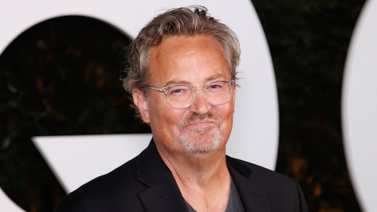 En mann i svart jakke og grå T-skjorte poserer foran en lys bakgrunn. Han har kort, bølgete hår. Bildet er tatt på et arrangement, med grønne planter synlige i bakgrunnen. Det er noe uklart i ansiktet hans.