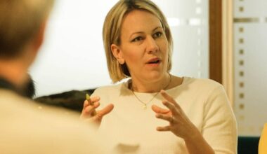 En kvinne i hvit genser gestikulerer med hendene under en samtale. Hun holder en penn i høyre hånd. Omgivelsene ser ut til å være et møterom med flere personer til stede. Det er en diskusjon i gang, og en annen person delvis synlig i bildet. (Bildebeskrivelsen er laget av en KI-tjeneste)