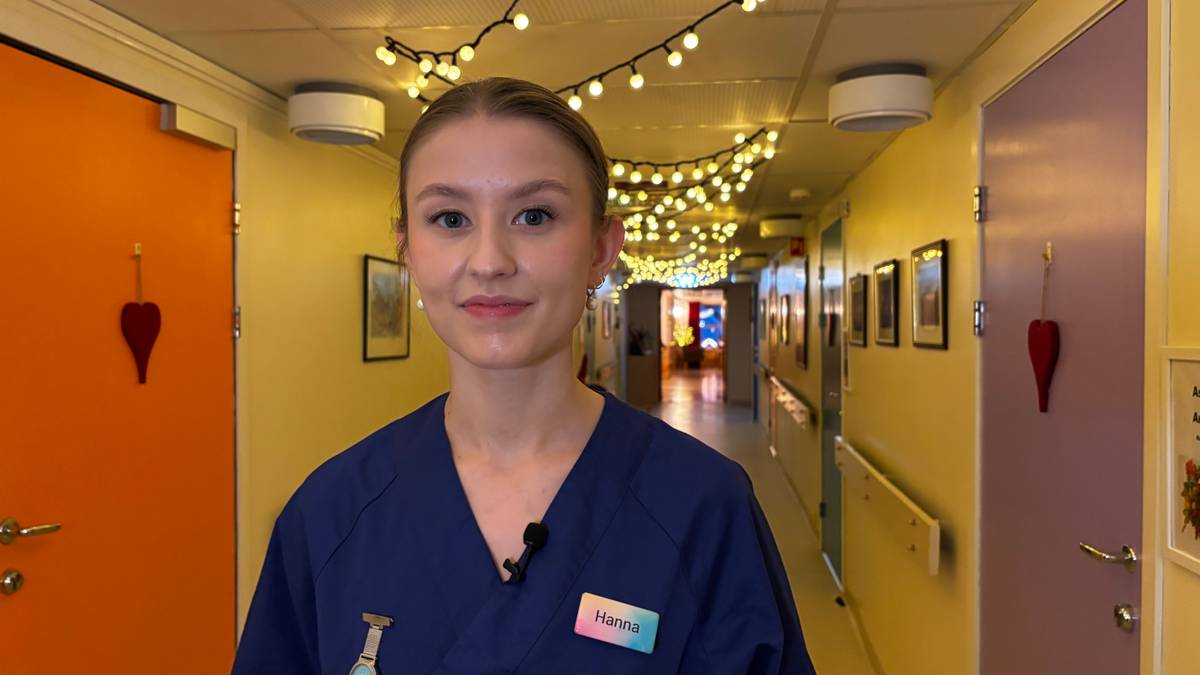 En person i blå arbeidsdrakt står i en gang med dekorative lys hengende fra taket. Bak personen er det dører i ulike farger, samt bilder på veggene. Belysningen gir en varm atmosfære i rommet. Det ser ut til å være et miljø hvor mennesker oppholder seg. (Bildebeskrivelsen er laget av en KI-tjeneste)