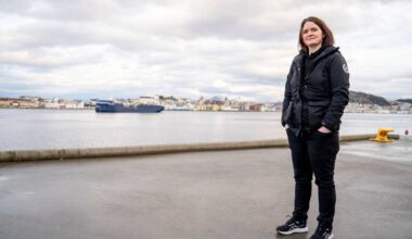 En person står på en kaiplass med havet og byen i bakgrunnen. Det er grå skyer over horisonten. En stor båt er synlig på vannet. Personen har på seg en svart jakke, bukser og sneakers.