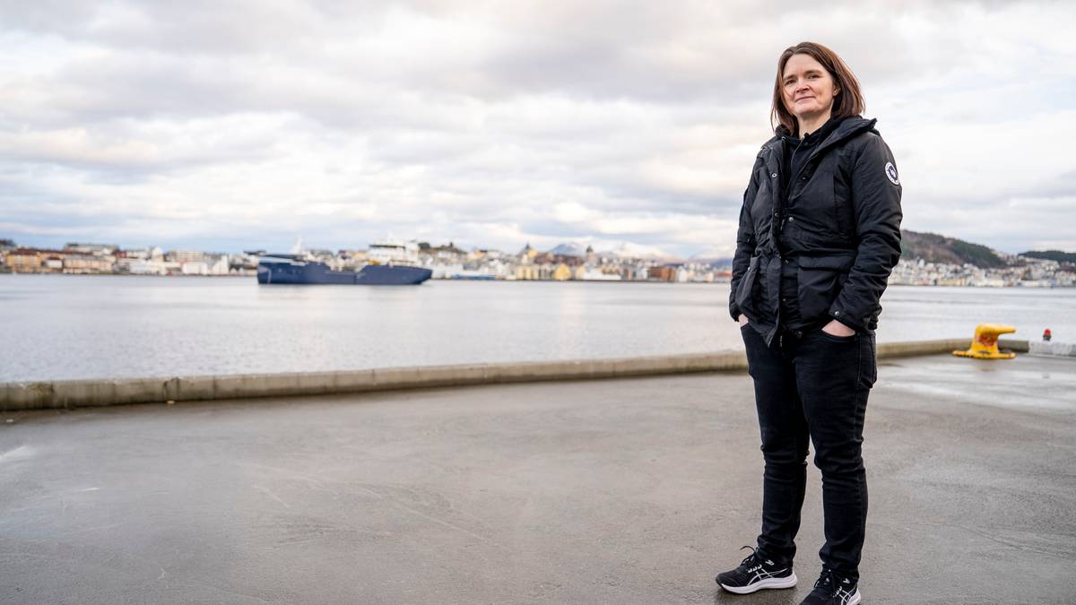 En person står på en kaiplass med havet og byen i bakgrunnen. Det er grå skyer over horisonten. En stor båt er synlig på vannet. Personen har på seg en svart jakke, bukser og sneakers.