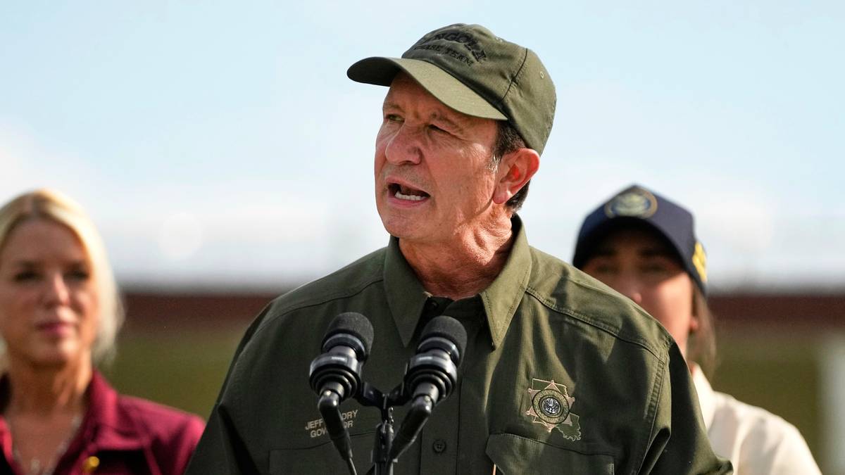 En mann står ved en talerstol under en pressekonferanse, iført grønn uniform og caps. Han holder en mikrofon og snakker, mens to andre personer står bak ham, iført offisielle klær. Talerstolen har et skilt med teksten "Louisiana". Bakgrunnen viser en utendørs scene, delvis uskarp. (Bildebeskrivelsen er laget av en KI-tjeneste)