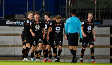 Fotballspillere feirer et mål sammen på banen. De har på seg svarte drakter med logoer. En dommer står vendt mot spillerne, klar til å gjenoppta spillet. Omgivelsene viser en stadion med gjerder og mørk belysning. (Bildebeskrivelsen er laget av en KI-tjeneste)
