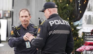 To polititjenestemenn i uniform snakker sammen utendørs. Det er juletre og dekorasjoner i bakgrunnen. En forbipasserende person står i nærheten. Begge politibetjentene har utstyr og merker synlig. (Bildebeskrivelsen er laget av en KI-tjeneste)