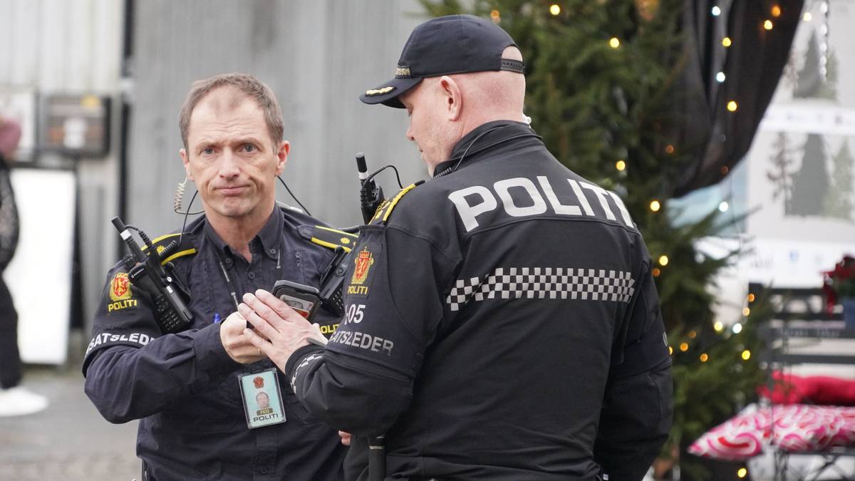 To polititjenestemenn i uniform snakker sammen utendørs. Det er juletre og dekorasjoner i bakgrunnen. En forbipasserende person står i nærheten. Begge politibetjentene har utstyr og merker synlig. (Bildebeskrivelsen er laget av en KI-tjeneste)