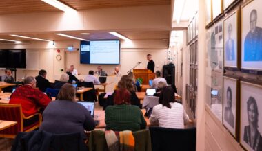 Et møte pågår i en romslig konferansesal med deltakerne samlet rundt bordene. En mann står ved en talerstol og presenterer, mens flere deltakere ser mot skjermen foran. Noen av deltakerne bruker bærbare datamaskiner, og en stor skjerm viser informasjon om møtet. Veggen er dekorert med innrammede bilder av tidligere deltakere, som bidrar til settingen. (Bildebeskrivelsen er laget av en KI-tjeneste)ne