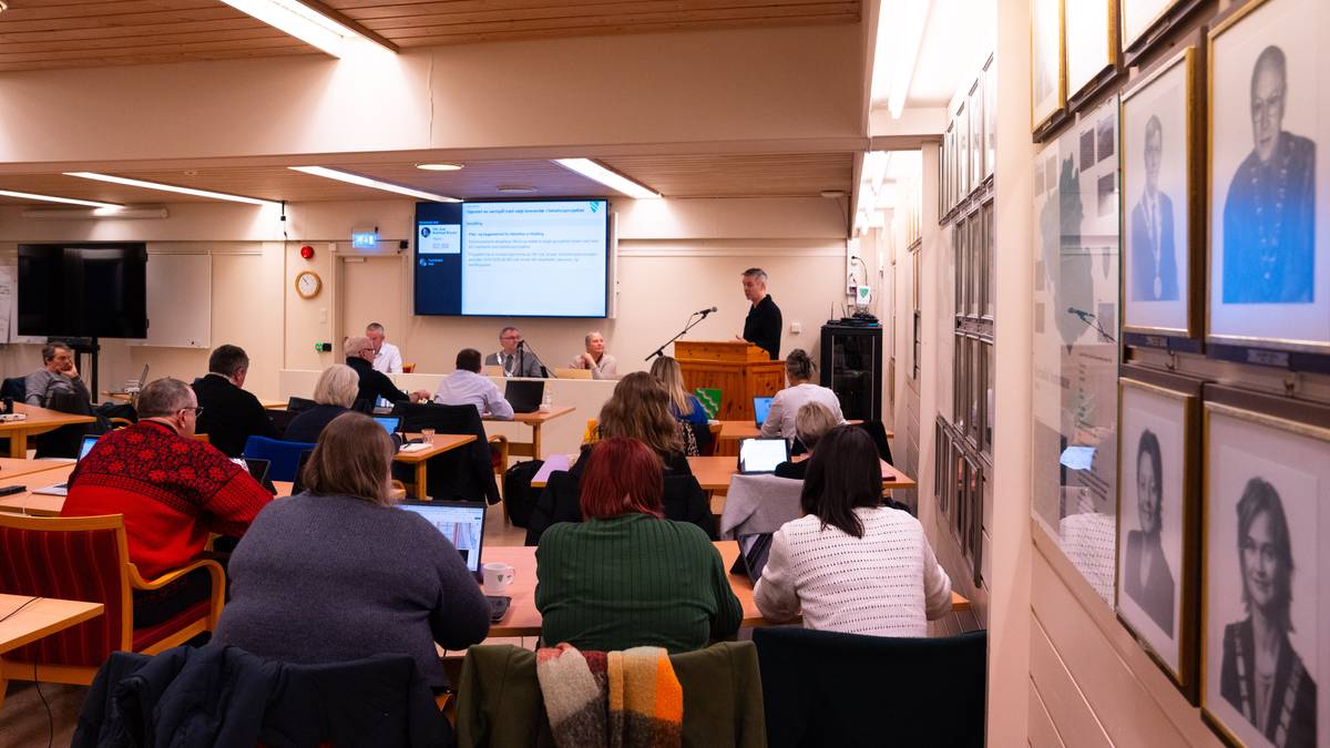 Et møte pågår i en romslig konferansesal med deltakerne samlet rundt bordene. En mann står ved en talerstol og presenterer, mens flere deltakere ser mot skjermen foran. Noen av deltakerne bruker bærbare datamaskiner, og en stor skjerm viser informasjon om møtet. Veggen er dekorert med innrammede bilder av tidligere deltakere, som bidrar til settingen. (Bildebeskrivelsen er laget av en KI-tjeneste)ne