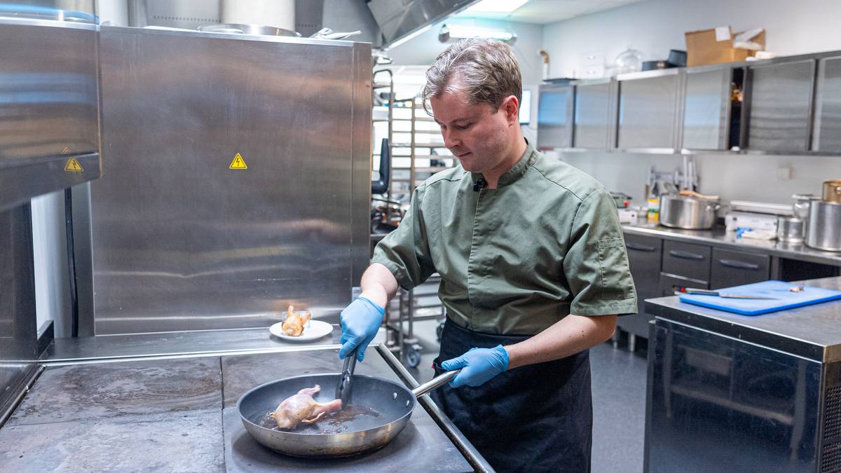 En kokk steker kyllinglår i en panne. Han bruker tenger og har på seg blå engangshansker. I bakgrunnen ses kjøkkenutstyr og en tallerken med mat. Kokken er iført en mørk grønn kokkeskjorte. (Bildebeskrivelsen er laget av en KI-tjeneste)