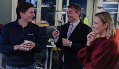 Tre personer står i et kontorlandskap og diskuterer. En av dem holder en pappkop, mens de andre gestikulerer. Det er datamaskiner og kontormøbler i bakgrunnen. Atmosfæren virker uformell. (Bildebeskrivelsen er laget av en KI-tjeneste)