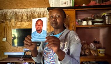 Ei afrikansk kvinne held opp eit fotografi av mannen sin. Også han er afrikansk. Ho står i eit rom, med ei litt rotete hylle bak seg. Ho ser inn i kamera.