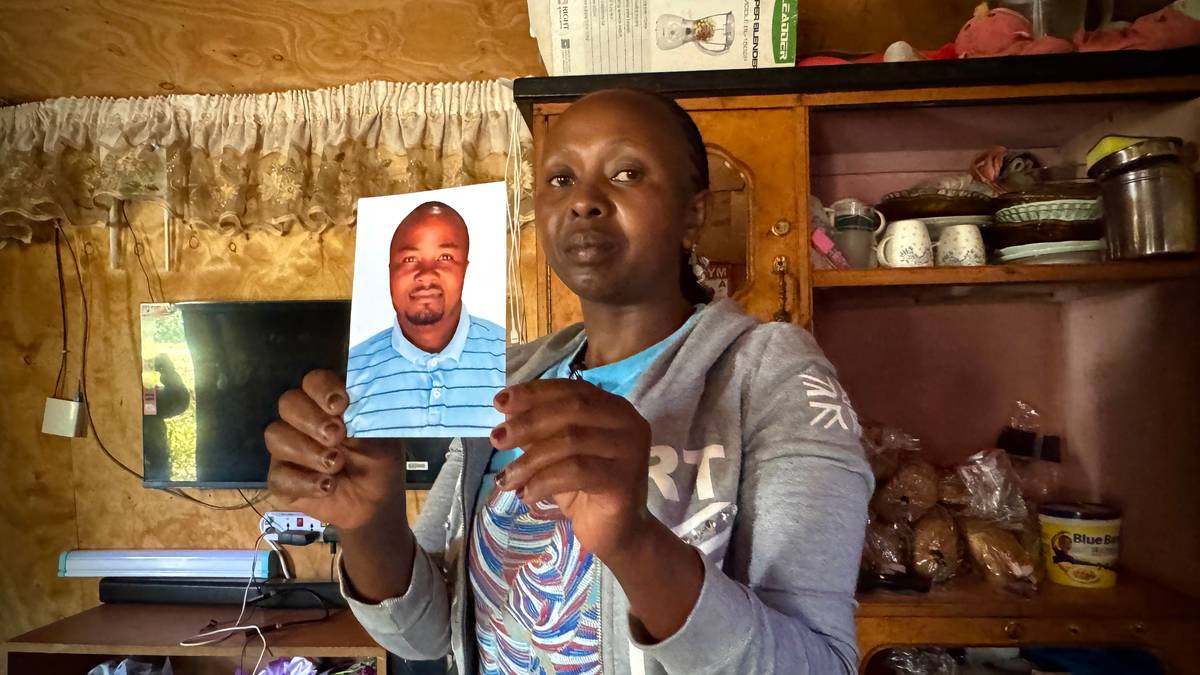 Ei afrikansk kvinne held opp eit fotografi av mannen sin. Også han er afrikansk. Ho står i eit rom, med ei litt rotete hylle bak seg. Ho ser inn i kamera.