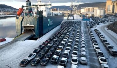 Et ferjeskip er ankommet havnen, der det ligger ved kai med rampen ned i snøen. Foran skipet står det i alt flere titalls biler parkert ryddig i rader. I bakgrunnen kan man se industriområder med kraner og bygninger langs elven. Landskapet er omkranset av åser og et klart vinterlys. (Bildebeskrivelsen er laget av en KI-tjeneste)