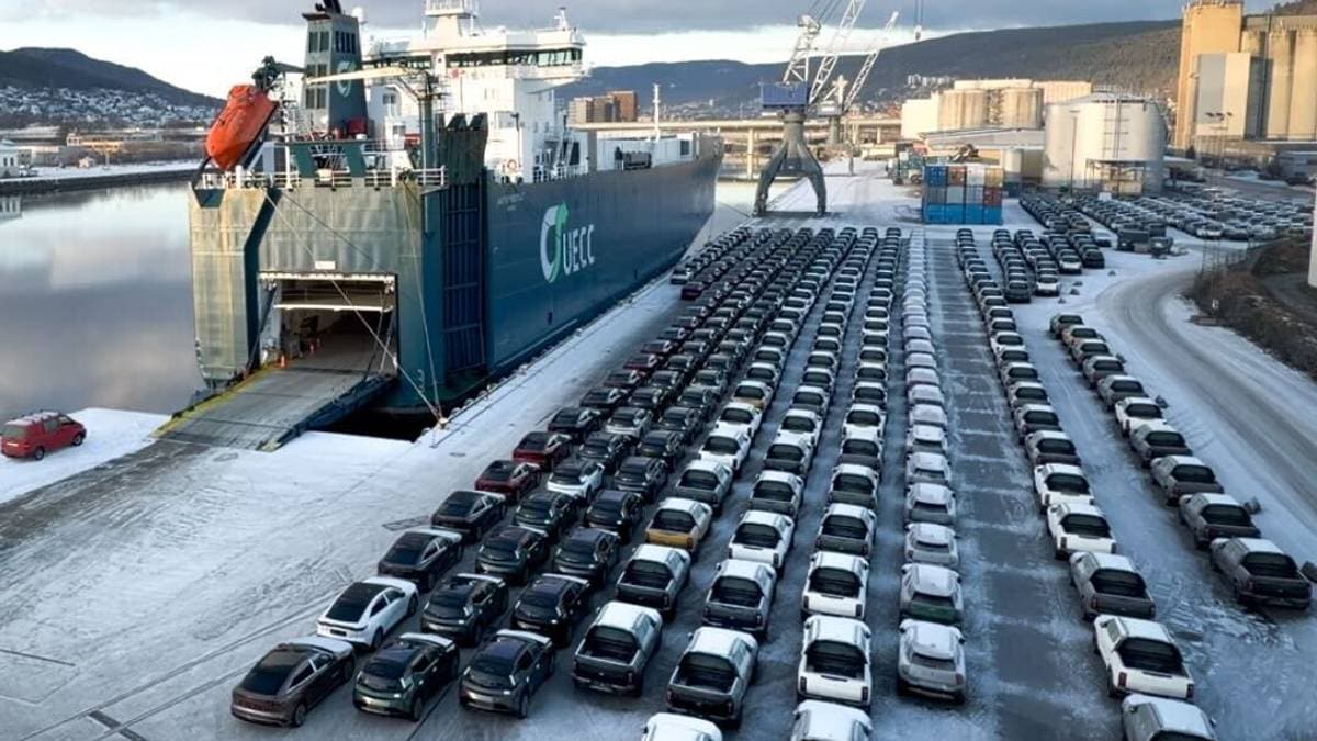 Et ferjeskip er ankommet havnen, der det ligger ved kai med rampen ned i snøen. Foran skipet står det i alt flere titalls biler parkert ryddig i rader. I bakgrunnen kan man se industriområder med kraner og bygninger langs elven. Landskapet er omkranset av åser og et klart vinterlys. (Bildebeskrivelsen er laget av en KI-tjeneste)