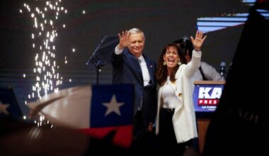 To personer står på en scene og vinkes til publikum under en arrangement. Rundt dem er det festlig dekor med gnister og flagg. En talerstol med teksten "KA PRES" er synlig foran dem. Stemningen virker høytidelig med nasjonale symboler i bakgrunnen. (Bildebeskrivelsen er laget av en KI-tjeneste)