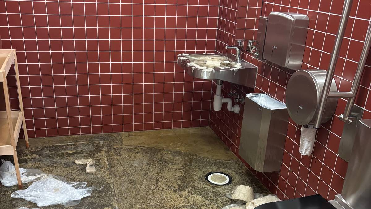 Bilde av et offentlig toalett med røde fliser på veggene. Gulvet er skittent og det ligger avfall, inkludert plastposer og papirruller, rundt. Toalettet er moderne og av rustfritt stål, med en vask og håndtørker synlig i rommet. Omgivelsene gir inntrykk av manglende vedlikehold. (Bildebeskrivelsen er laget av en KI-tjeneste)