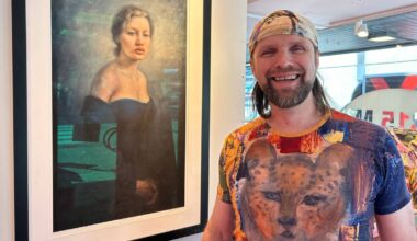 En person står foran et innrammet maleri av en kvinne iført en blå kjole. Personen har på seg en fargerik T-skjorte med dyremotiv. Omgivelsene inkluderer et moderne galleri med naturlig lys som strømmer inn gjennom vinduer. Refleksjoner på glasset bidrar til å skape en hendelse i rommet. (Bildebeskrivelsen er laget av en KI-tjeneste)Storfornøyd Aune Sand