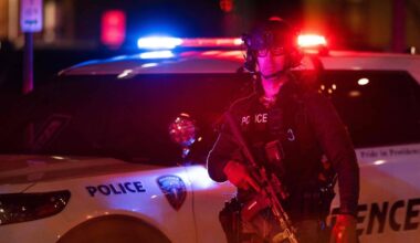 En politibetjent i taktisk utstyr står foran en politibil med blått og rødt lys aktivert. Betjenten holder et automatvåpen mens han overvåker situasjonen. Politibilen har tydelige politimerker og står parkert langs en gate om natten. Omgivelsene er svakt belyst, fokus er sentrert på politiets tilstedeværelse. (Bildebeskrivelsen er laget av en KI-tjeneste)