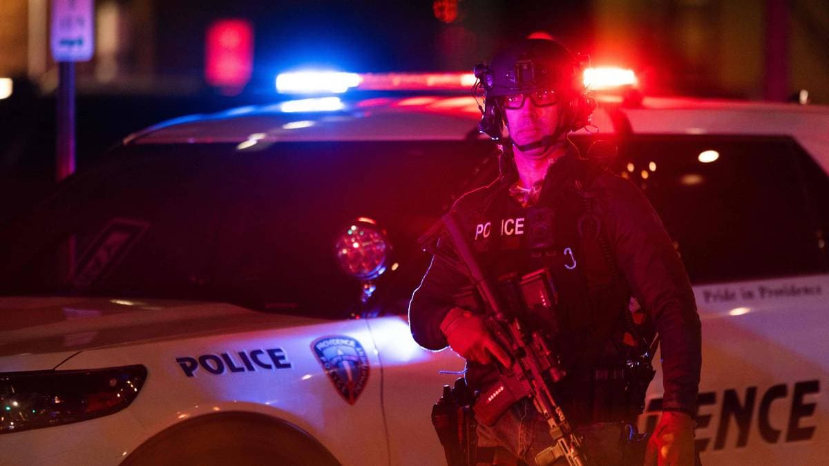 En politibetjent i taktisk utstyr står foran en politibil med blått og rødt lys aktivert. Betjenten holder et automatvåpen mens han overvåker situasjonen. Politibilen har tydelige politimerker og står parkert langs en gate om natten. Omgivelsene er svakt belyst, fokus er sentrert på politiets tilstedeværelse. (Bildebeskrivelsen er laget av en KI-tjeneste)