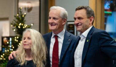 Tre personer står sammen foran et industrielt bakteppe. To av dem er kledd i mørke dresser, mens den ene har på seg en svart topp. Det er en juletre med lys i bakgrunnen, som gir en festlig stemning. Alle smiler mot kamera.