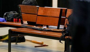 En benk i en offentlig bygning viser ulike eiendeler, inkludert en rifle, utstyr og ryggsekker. Det er også en baseballkølle i forgrunnen. Omgivelsene har et moderne interiør med fliser på gulvet og en dør i bakgrunnen. Ingen personer er synlige i bildet. (Bildebeskrivelsen er laget av en KI-tjeneste)