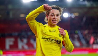 En fotballspiller i gult drakt hever hånden i jubel etter en kamp. Bakgrunnen viser en delvis fylt stadion. Spilleren ser ut til å feire et mål eller seier. Lysene på stadion er tent, og atmosfæren virker intens.