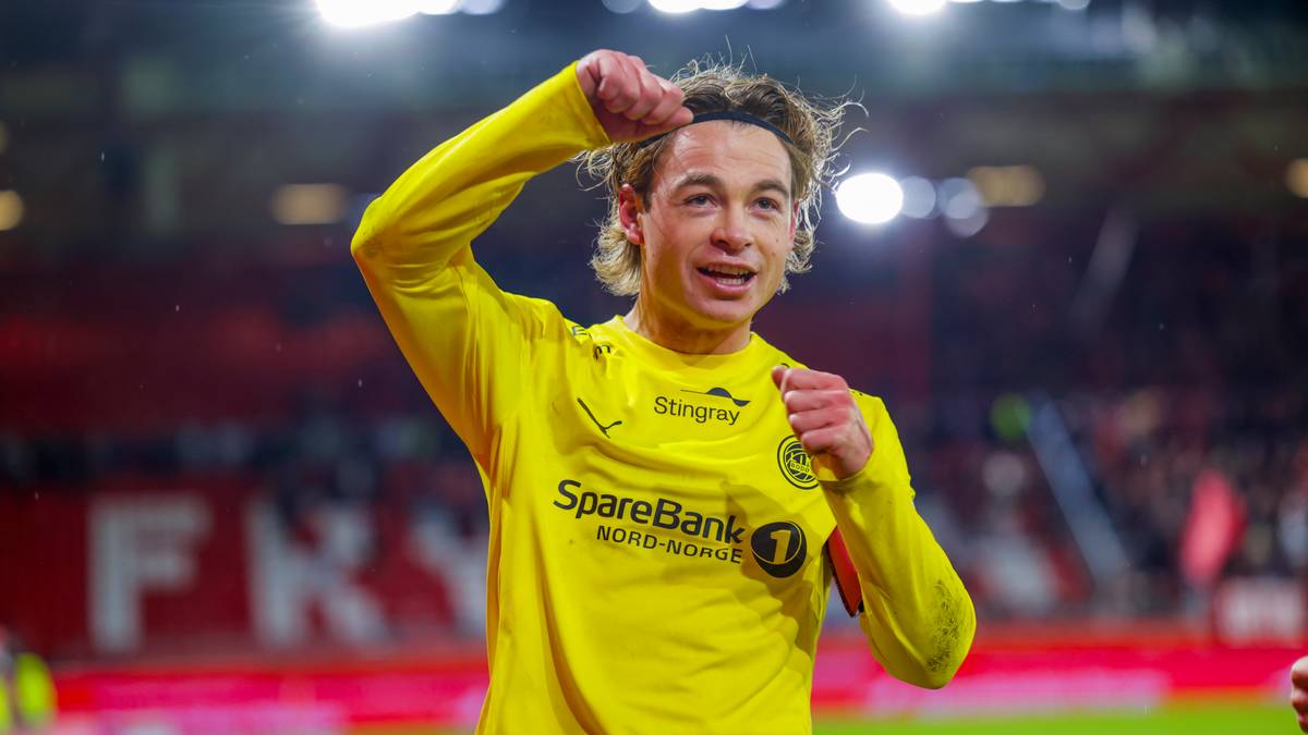 En fotballspiller i gult drakt hever hånden i jubel etter en kamp. Bakgrunnen viser en delvis fylt stadion. Spilleren ser ut til å feire et mål eller seier. Lysene på stadion er tent, og atmosfæren virker intens.