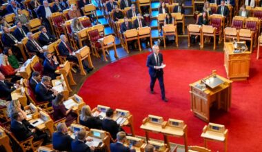 En person står foran en forsamling i en stortingssal, og holder et innlegg. Publikum består av mange personer i formelle klær, både menn og kvinner, som følger med. Rød teppe dekker gulvet, og det er flere skrivebord med digitale enheter foran deltakerne. Bakgrunnen viser et rom med treplanker og stoler i tradisjonell stil.