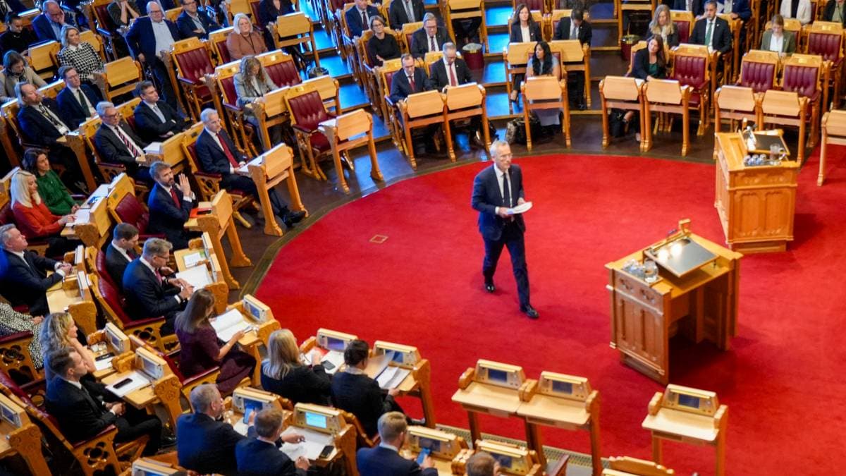 En person står foran en forsamling i en stortingssal, og holder et innlegg. Publikum består av mange personer i formelle klær, både menn og kvinner, som følger med. Rød teppe dekker gulvet, og det er flere skrivebord med digitale enheter foran deltakerne. Bakgrunnen viser et rom med treplanker og stoler i tradisjonell stil.