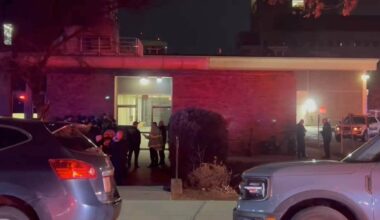 Politi og redningspersonell samles utenfor en bygning om natten. Flere personer står i grupper og diskuterer. Det er utrykningskjøretøy parkert i nærheten, og bygningen er opplyst av lys fra inngangen. Omgivelsene er preget av mørke, med noen lys fra nærliggende bygninger. (Bildebeskrivelsen er laget av en KI-tjeneste)