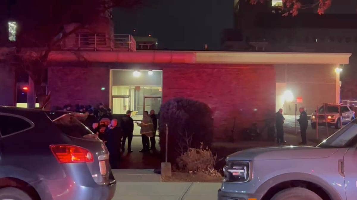 Politi og redningspersonell samles utenfor en bygning om natten. Flere personer står i grupper og diskuterer. Det er utrykningskjøretøy parkert i nærheten, og bygningen er opplyst av lys fra inngangen. Omgivelsene er preget av mørke, med noen lys fra nærliggende bygninger. (Bildebeskrivelsen er laget av en KI-tjeneste)